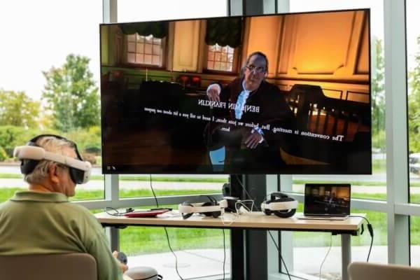 An individual wears a virtual reality headset as a TV displays a virtual avatar of Benjamin Franklin in Independence Hall.